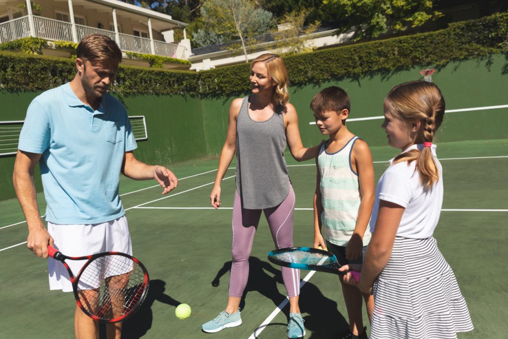 Family standing on a tennis court, with a father holding a tennis racket and giving instructions, mom looks at dad with a smile. 